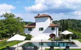 Elegant villa with a swimming pool surrounded by sun loungers and umbrellas, set against a lush green landscape and blue sky. Ideal for luxury retreats or vacation rentals in a scenic location.