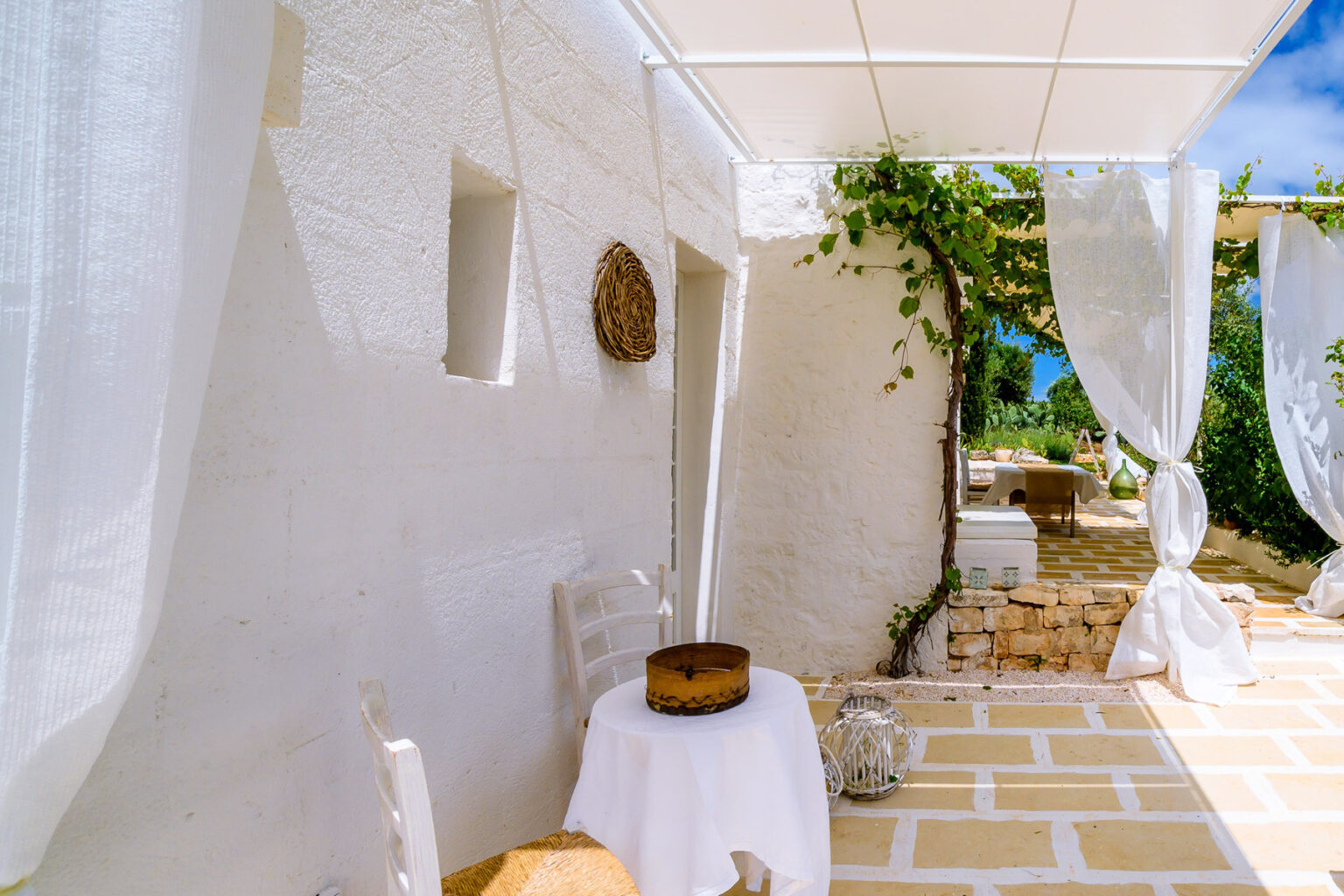 Cozy outdoor terrace with white walls, a round table set for dining, a decorative cake, and flowing white curtains, surrounded by greenery and stone pathways under a bright blue sky. | Only on SopranoVillas