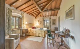 Cozy bedroom with wooden beams, featuring a large bed, comfortable seating area, and a desk, surrounded by natural light from windows overlooking a scenic view.