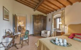 Cozy bedroom featuring wooden ceiling beams, a vintage wardrobe, a comfortable armchair, and a small writing desk, all adorned with warm colors and elegant decor. A tray with tea set adds a touch of hospitality.