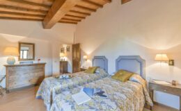 Cozy guest room with twin beds, floral bedding, wooden beams, and warm lighting, featuring a wooden dresser and a mirror, ideal for a relaxing stay.