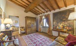 Cozy living room featuring wooden beam ceilings, elegant furniture, and a large floral painting. The space includes a comfortable seating area, a decorative rug, and warm lighting, creating an inviting atmosphere.