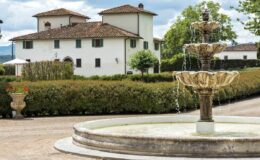 Elegant villa with a classic Italian architectural style, featuring a central fountain surrounded by manicured hedges and lush greenery. The serene landscape is complemented by a clear sky, creating a picturesque setting for relaxation and leisure.