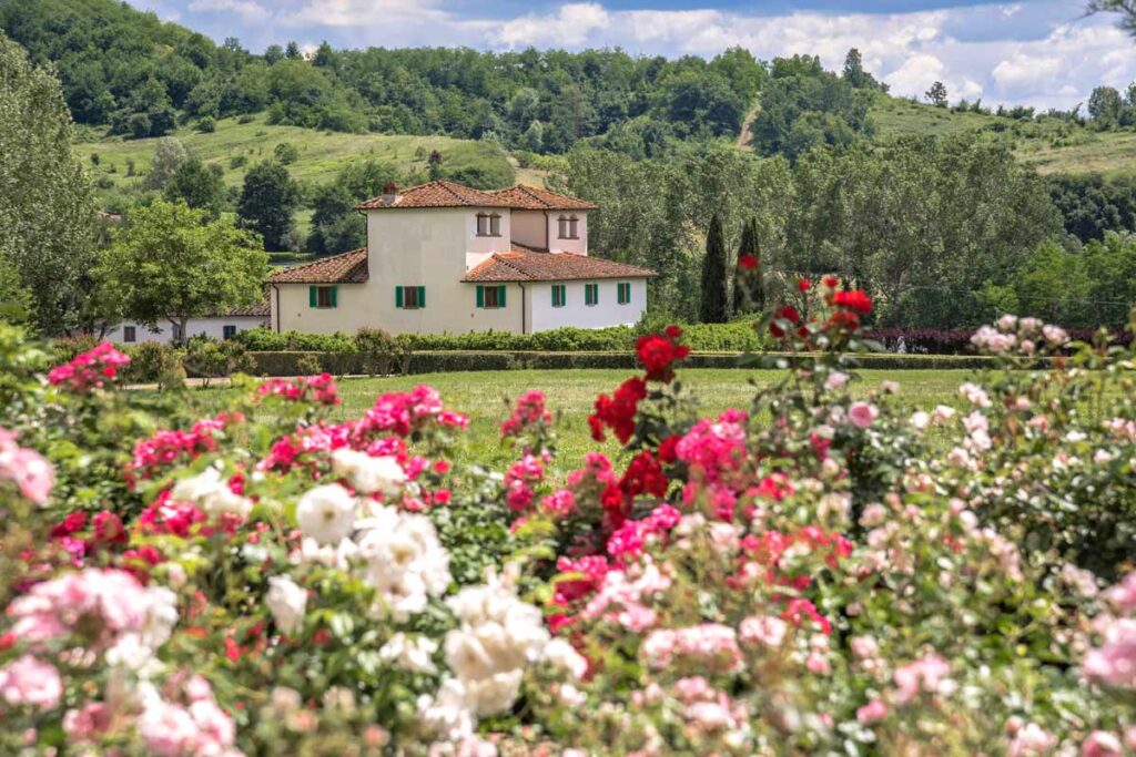 Beautiful countryside house surrounded by vibrant rose gardens, with lush green hills in the background under a clear blue sky. | Only on SopranoVillas