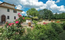 Scenic view of a charming Italian villa surrounded by vibrant rose bushes and lush greenery under a clear blue sky with fluffy clouds.