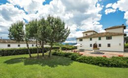 Elegant Italian villa surrounded by lush greenery and blue skies, featuring manicured lawns, trees, and a patio area. Ideal for outdoor gatherings and relaxation.