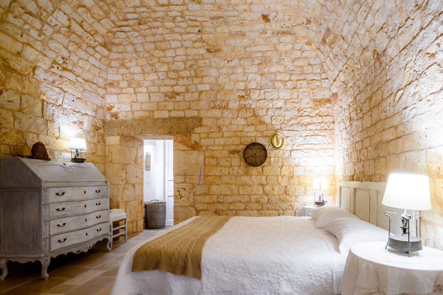 Cozy bedroom with rustic stone walls, featuring a white bed with a beige throw, vintage dresser, and warm lighting from bedside lamps, creating a welcoming atmosphere. | Only on SopranoVillas