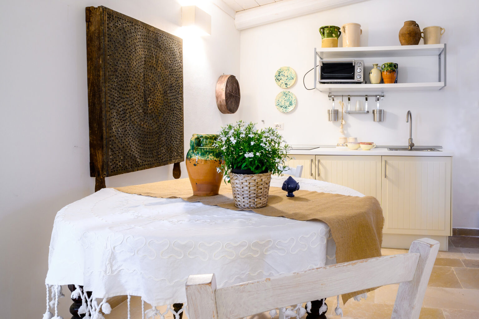 Cozy kitchen interior featuring a round table with a white tablecloth, potted plants, and decorative wall art. The kitchen area includes light cabinetry, a microwave, and simple kitchenware, creating a warm and inviting atmosphere. | Only on SopranoVillas