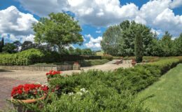 Lush garden landscape featuring well-maintained hedges, vibrant red flowers in planters, and a scenic pathway under a blue sky with fluffy clouds.