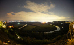 Panoramic night view of a river winding through lush landscapes, illuminated by city lights in the background, showcasing a serene and picturesque natural setting.