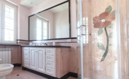 Elegant bathroom featuring a large mirror, white cabinetry, and a glass shower with floral design. Natural light streams through windows, highlighting the pink marble walls and black accents.