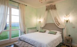 Elegant bedroom with a light green color scheme, featuring a plush bed adorned with decorative pillows, soft lighting from wall sconces, and large windows that offer a view of the outdoors. The room is designed for comfort and relaxation, with tasteful decor and a cozy atmosphere.