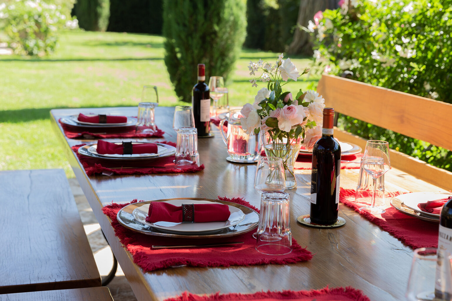 Elegant outdoor dining setup featuring a long wooden table adorned with red table runners, plates, glasses, and wine bottles, surrounded by lush greenery. Perfect for a summer gathering or celebration. | Only on SopranoVillas