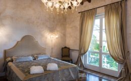 Elegant bedroom featuring a plush bed with decorative pillows, a luxurious chandelier, and large windows draped with golden curtains, creating a warm and inviting atmosphere.