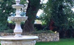 Stone garden fountain surrounded by lush greenery and trees, with a rustic stone wall in the background.