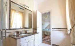 Bright and airy bathroom featuring a large mirror, white cabinetry, and a marble countertop, with natural light streaming through sheer curtains. The doorway leads to a bedroom, enhancing the spacious feel of the area.