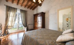 Cozy bedroom featuring a queen-sized bed with a neutral-colored quilt, wooden flooring, and large windows with drapes, allowing natural light to enter. The room includes a vintage wooden wardrobe and a stylish mirror, creating a warm and inviting atmosphere.