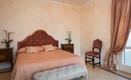 Cozy bedroom featuring a pink upholstered bed with decorative pillows, a stylish bench at the foot, and elegant wooden nightstands. Soft peach walls and natural light enhance the inviting atmosphere, complemented by a vintage chair and floral accents.