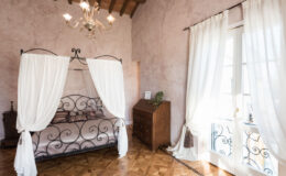 Elegant bedroom featuring a wrought iron four-poster bed with white drapes, a wooden dresser, and large windows adorned with sheer curtains, creating a bright and airy atmosphere.
