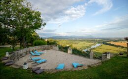 Scenic outdoor lounge area with turquoise sun loungers overlooking a lush valley and river, surrounded by greenery and a dramatic sky. Ideal for relaxation and enjoying nature.