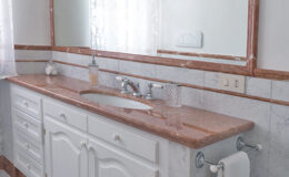 Modern bathroom vanity with a pink marble countertop, white cabinetry, and a large mirror, featuring natural light from a nearby window.
