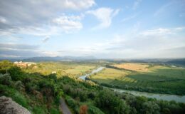 Panoramic view of a lush green landscape with a winding river, rolling hills, and a blue sky dotted with clouds, showcasing the beauty of nature.