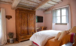 Cozy bedroom featuring a wooden wardrobe, a comfortable bed with decorative pillows, and natural light streaming through window curtains, set against a pink wall and rustic wooden ceiling.