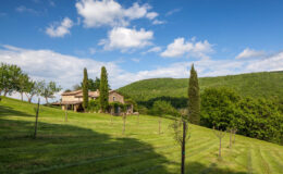 Scenic view of a rustic farmhouse surrounded by lush green hills and neatly manicured lawns under a clear blue sky in a picturesque countryside setting.