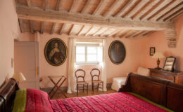 Cozy bedroom interior featuring a wooden bed with a vibrant red quilt, vintage portraits on the walls, rustic wooden ceiling beams, and a small seating area with two chairs. Natural light streams through the window, enhancing the warm ambiance of the room.
