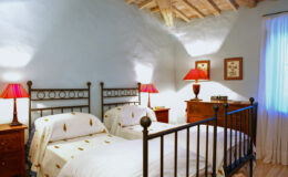 Cozy bedroom featuring two twin beds with decorative bedspreads, wooden furniture, and warm red lamps, set against a light blue wall and a wooden ceiling. Ideal for a comfortable stay.