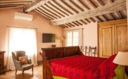 Cozy bedroom featuring a wooden bed with a red and green bedspread, rustic wooden beams on the ceiling, a comfortable armchair, a flat-screen TV, and natural light from large windows. Ideal for a relaxing getaway.