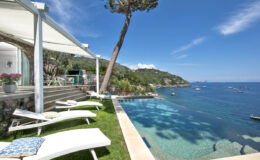 Luxury villa with infinity pool overlooking the Mediterranean Sea, surrounded by lush greenery and a clear blue sky, featuring sun loungers and a modern architectural design.