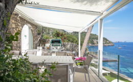 Cozy outdoor dining area with a view of the sea, featuring a stone wall, white furniture, and vibrant flowers, set under a retractable awning. Ideal for enjoying meals in a scenic coastal environment.