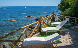 Lounge chairs on a wooden deck overlooking a clear blue sea, with rocky shoreline and boats in the distance, offering a serene coastal retreat.