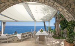 Elegant outdoor dining area under a retractable awning, featuring modern furniture and a stunning ocean view, surrounded by lush greenery and stone architecture. Perfect for alfresco gatherings and relaxation.