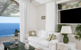 Bright and airy coastal living room with a white sofa, decorative pillows, and a green side table, featuring large windows that offer a stunning ocean view.