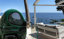 Luxury beachfront terrace with modern seating, overlooking a calm ocean view and a green outdoor pod, surrounded by trees.