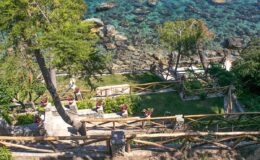Scenic coastal view featuring a winding pathway through lush greenery, leading down to clear turquoise waters and rocky shoreline, with visitors enjoying the natural beauty.