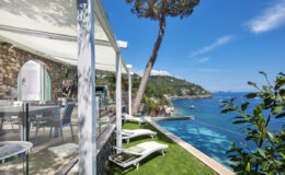 Luxury seaside villa with modern outdoor seating, infinity pool, and stunning ocean views, surrounded by lush greenery and a clear blue sky.