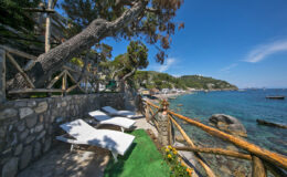 Coastal view featuring sun loungers beside a stone pathway, surrounded by lush greenery and clear blue waters, ideal for relaxation and enjoying nature.