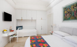 Bright and modern bedroom featuring a double bed, a single bed, a wall-mounted TV, and a decorative painting. The room is adorned with a colorful floral bedspread and has minimalist white furniture, creating a spacious and inviting atmosphere.
