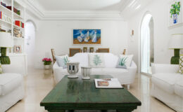 Bright and airy living room featuring white sofas with green decorative pillows, a dark green coffee table, and a colorful painting on the wall. Shelves filled with books and decorative items add character, while natural light floods the space through arched doorways.