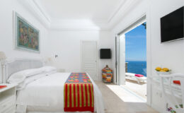 Bright and airy bedroom with a white color scheme, featuring a comfortable bed with colorful bedding, a flat-screen TV, and a door leading to a stunning ocean view. Decorated with art and a vibrant vase, the room exudes a relaxing coastal vibe.
