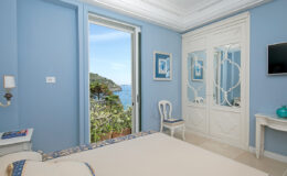 Cozy bedroom with blue walls, featuring a bed, elegant chair, and a wardrobe. Large open door leads to a scenic view of the ocean and greenery, enhancing the tranquil atmosphere. Modern decor includes a wall-mounted TV and decorative accents.