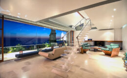 Modern luxury living room with a sleek glass staircase, contemporary furniture, and stunning ocean views at dusk. Natural light floods the space, highlighting the elegant design and open layout.