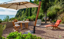 Cozy outdoor seating area with a wooden table and chairs under a large umbrella, overlooking a scenic coastal view. A lounge chair and hammock are nearby, surrounded by lush greenery and gravel landscaping, perfect for relaxation.