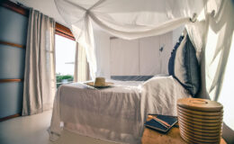 Cozy bedroom with a white canopy bed, soft lighting, and natural decor elements. Features a wooden side table with stacked books and a hat, creating a serene and inviting atmosphere.