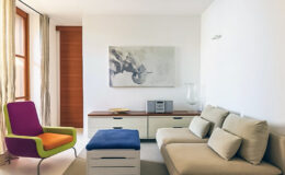 Modern living room featuring a beige sofa, colorful accent chair, and minimalist decor. A wooden cabinet holds a stereo system, while a large artwork hangs on the wall, enhancing the bright and airy atmosphere. Natural light filters through sheer curtains.