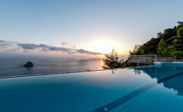 Infinity pool overlooking a serene ocean at sunset, surrounded by lush greenery and dramatic clouds.
