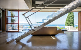 Modern interior featuring a sleek glass staircase leading to a panoramic view of the ocean, with large windows and minimalist decor.